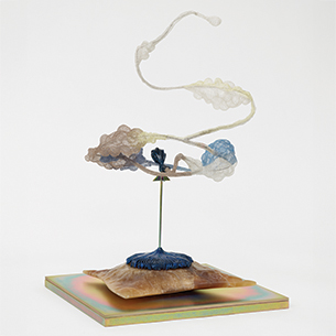 A sculptural installation featuring a central, translucent form with flowing, organic tendrils. The sculpture is mounted on a base with a colorful, iridescent surface.